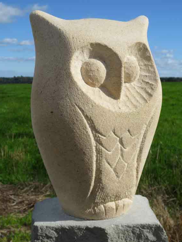 Eine Gartenskulptur aus Stein in Form einer Eule, umgeben von Blumen und Sträuchern