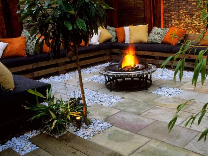 runde Feuerstelle aus Metall Gartengestaltung mit Steinen und Holz lange Sitzbank bunte Kissen