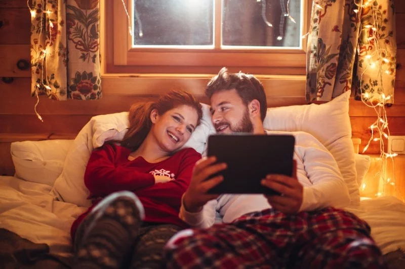 Couple lying on cozy bed in wooden cabin. Wearing Christmas pajamas. Together watching movie on digital tablet. Enjoying in the warmth of their home for Christmas. Room is decorated with festive string lights. Austrian Alps. Evening or night with beautiful yellow lights lightning the scenes.