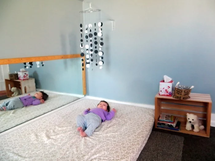 großes Montessori Bett mit Spiegel und kleines Baby beobachtet die hängende Deko 