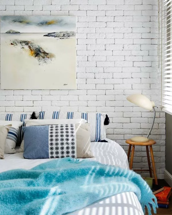 Backsteinwand im Schlafzimmer in Weiß gestrichen betont die ruhige Atmosphäre rustikales Flair Wandbild Hocker Nachtlampe