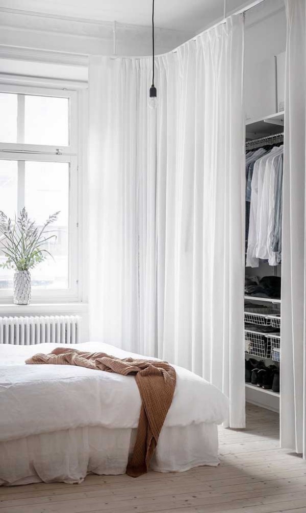 offener Kleiderschrank hinter feinen Gardinen im weißen Schlafzimmer