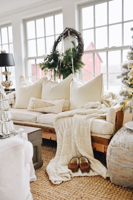 Winterdeko im Wohnzimmer ganz in Weiß gestaltet Strickdecke Kissen weißes Sofa rustikales Flair Kranz aus grünen Tannenzweigen am Fenster