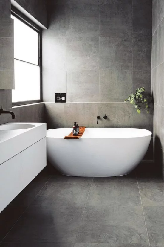 Baddesigns in Grau Minimalismus pur graue Boden-und Wandfliesen weiße Badewanne als Blickfang weißer Waschtisch Fenster rechts