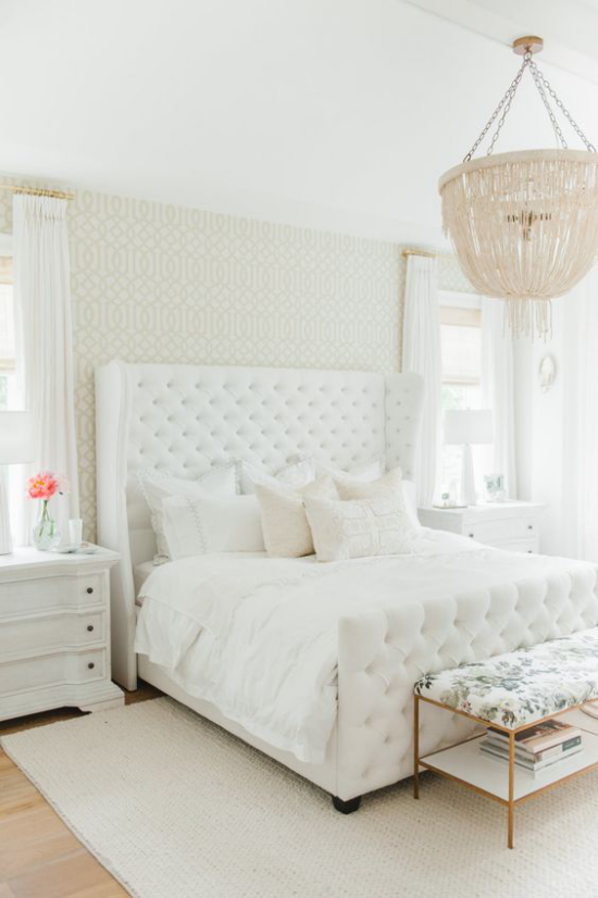 Glamouröse Schlafzimmer ganz in Weiß gestaltet gepolstertes Bett weiche Bettwäsche Deko Kissen Kronleuchter rosa Blume in Vase aus dem Nachttisch