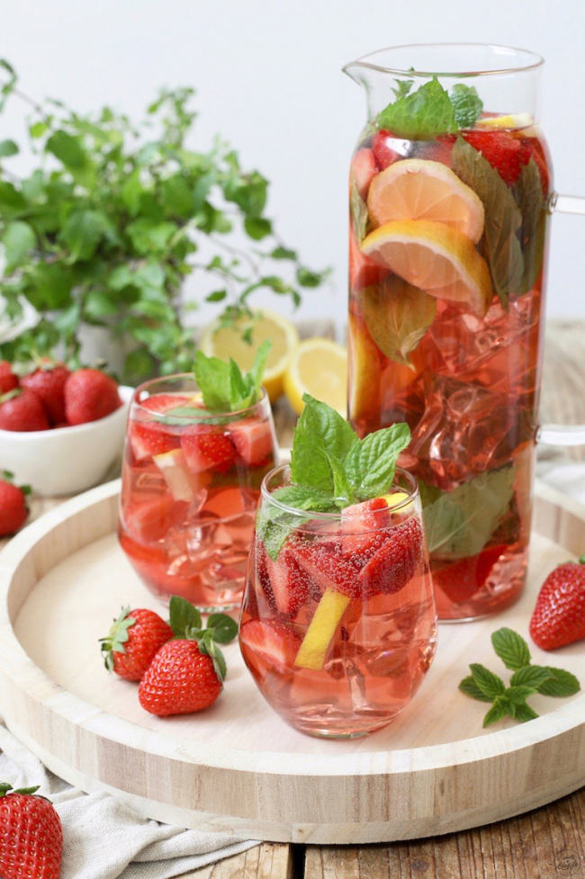 Sommerbowle zubereiten frisch lecker fruchtig Erdbeeren Zitronenscheiben Saft Blätter frische Minze in hohem Glasgefäß Gläser