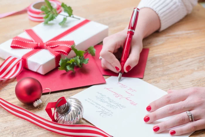 young woman writing christmas cards with red nails, a red pen, and holiday decorations