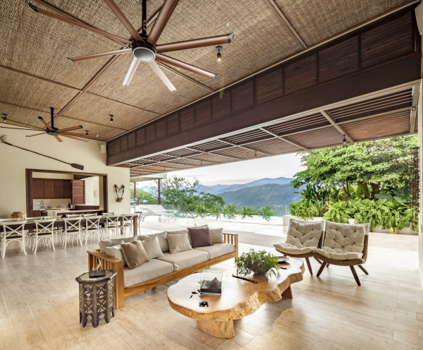 Open-Air-Wohnzimmer großzügiges Raumkonzept Blickfang Kaffeetisch aus Holz zwei Sessel bequemes Sofa im Hintergrund Esstisch viele Stühle