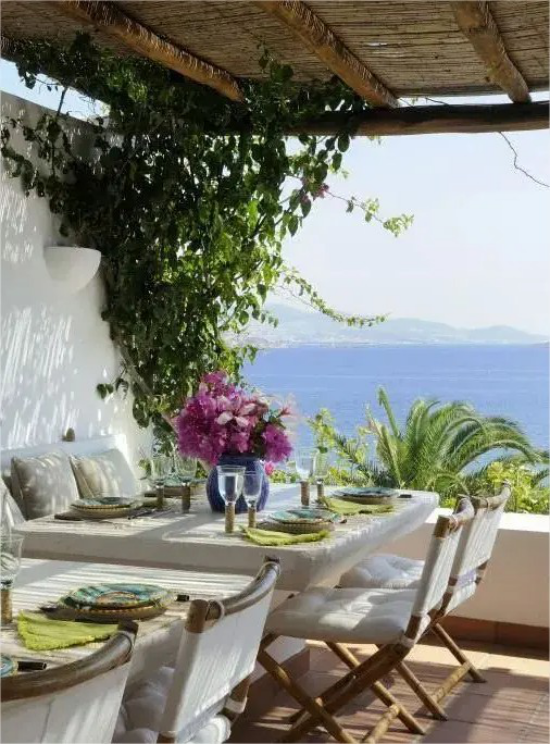 mediterraner Außenbereich überdachte Terrasse bedeckte Esstische weiße Tischdecke Korbgeflecht Vase mit Blumen Azurblau