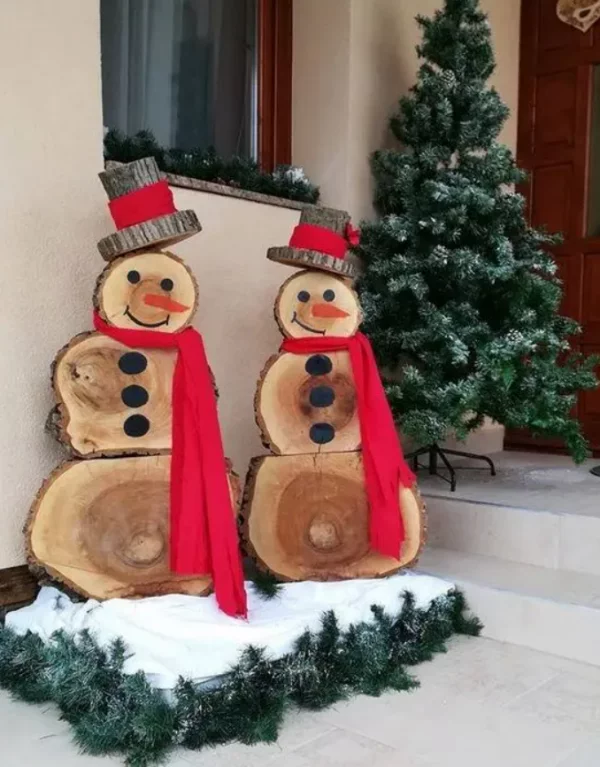 Schneemänner basteln lustige Weihnachtsdeko vor der haustuer zwei aus Holzscheiben mit roren Schals gruene Tannenzweige auf dem Boden